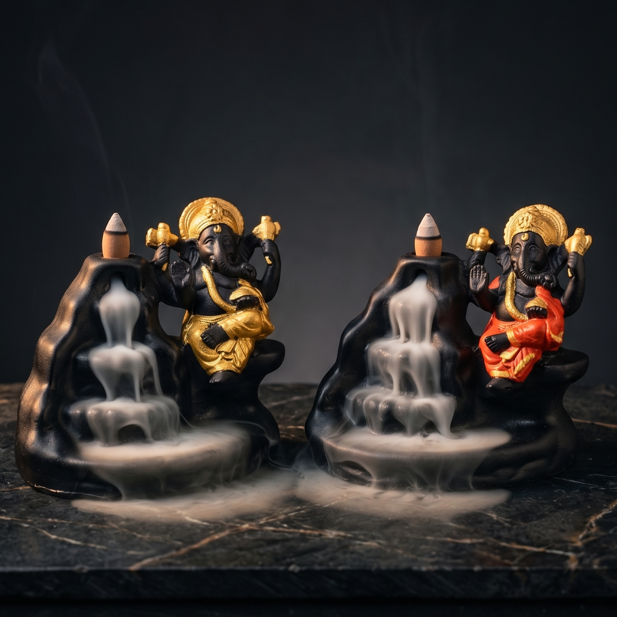 Two decorative incense burners shaped like seated Ganesha figures on black rock forms with cascading water, displayed on a dark marble surface against a black background. One figure is gold and the other is red, with smoke rising from the incense burners.