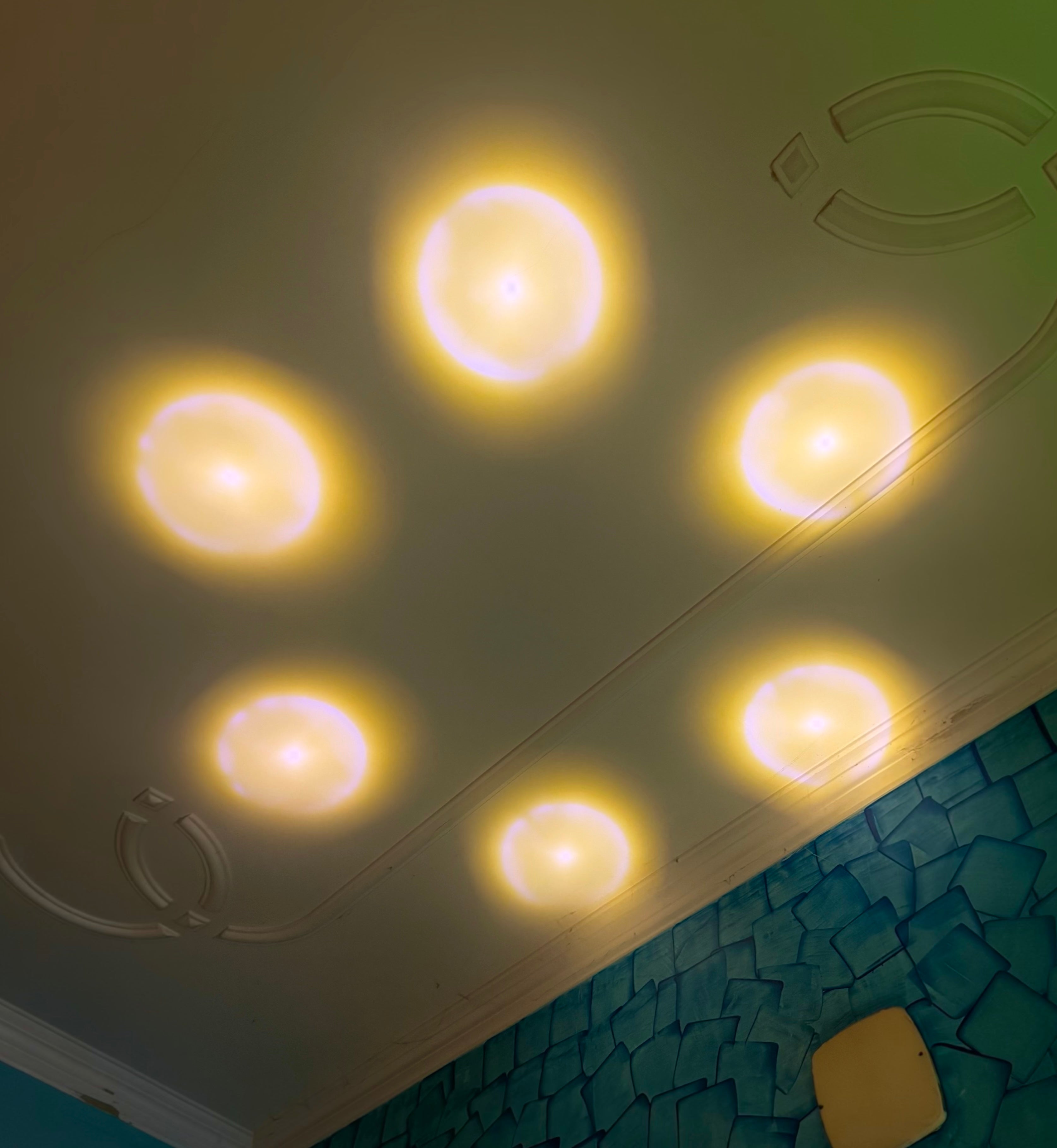 Radha Krishna 3D Crystal Night Lamp throwing beautiful uplight to the ceiling. Six round recessed ceiling lights glowing with a warm yellow light, installed on a white ceiling above a textured teal wall.