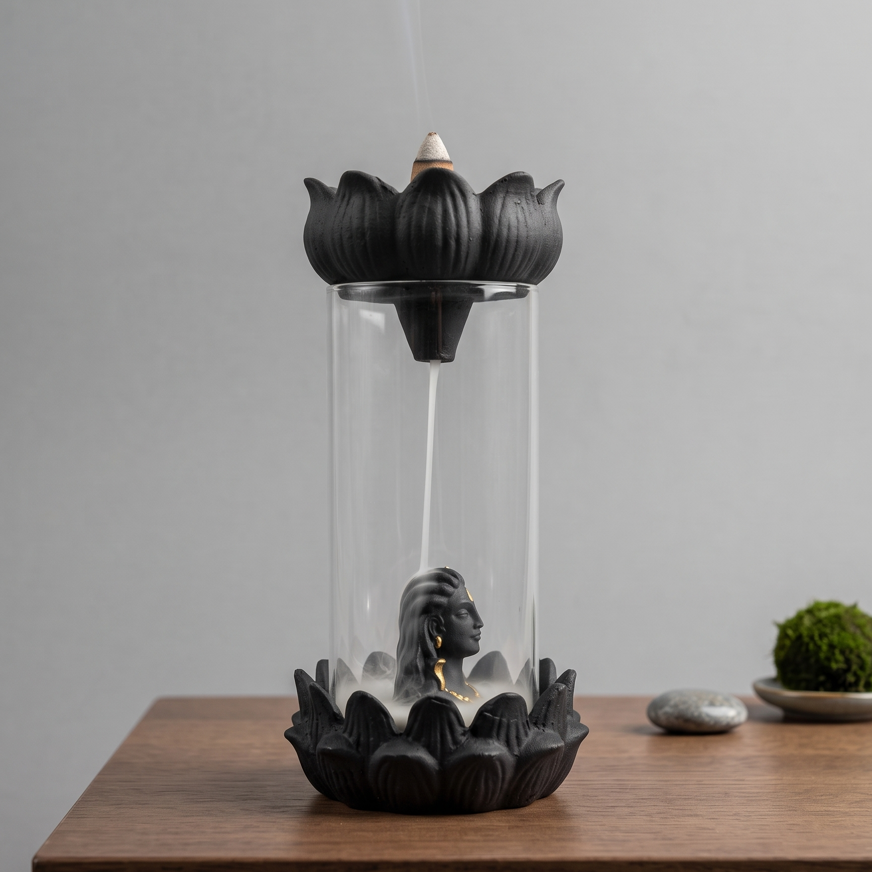 A black lotus-shaped incense burner with a clear cylindrical glass body sits on a wooden table against a light gray wall. A smoking incense stick is visible inside, with a small mossy stone and a gray decorative object in the background.